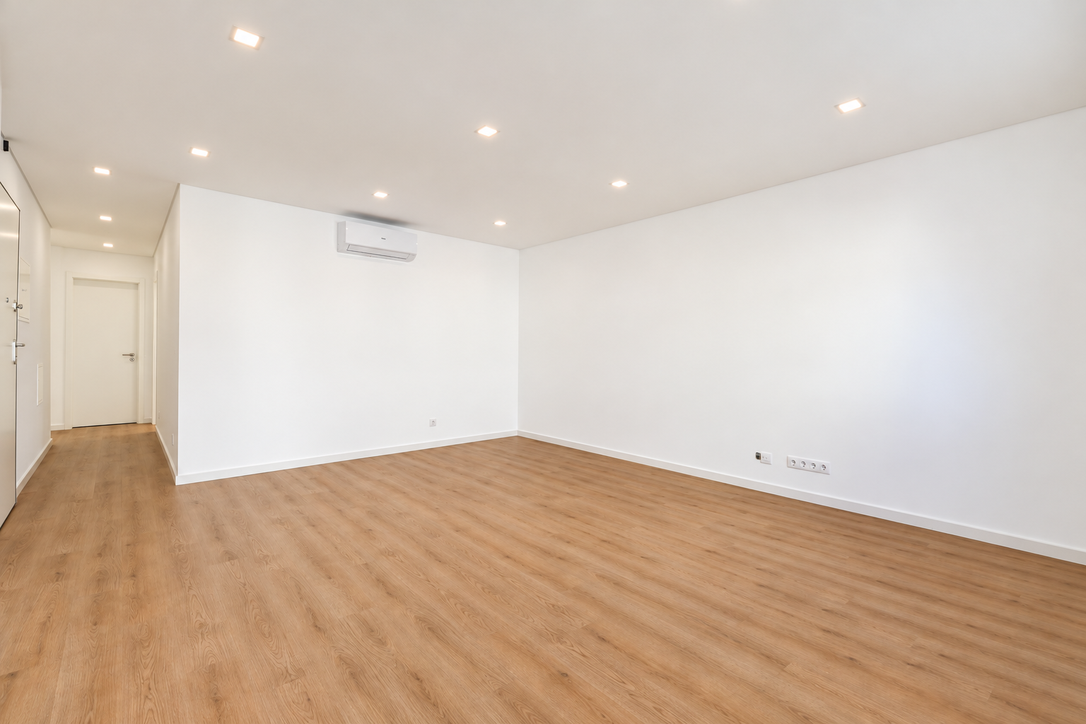 Parede branca da sala com ar condicionado, tomadas baixas e pavimento contínuo.