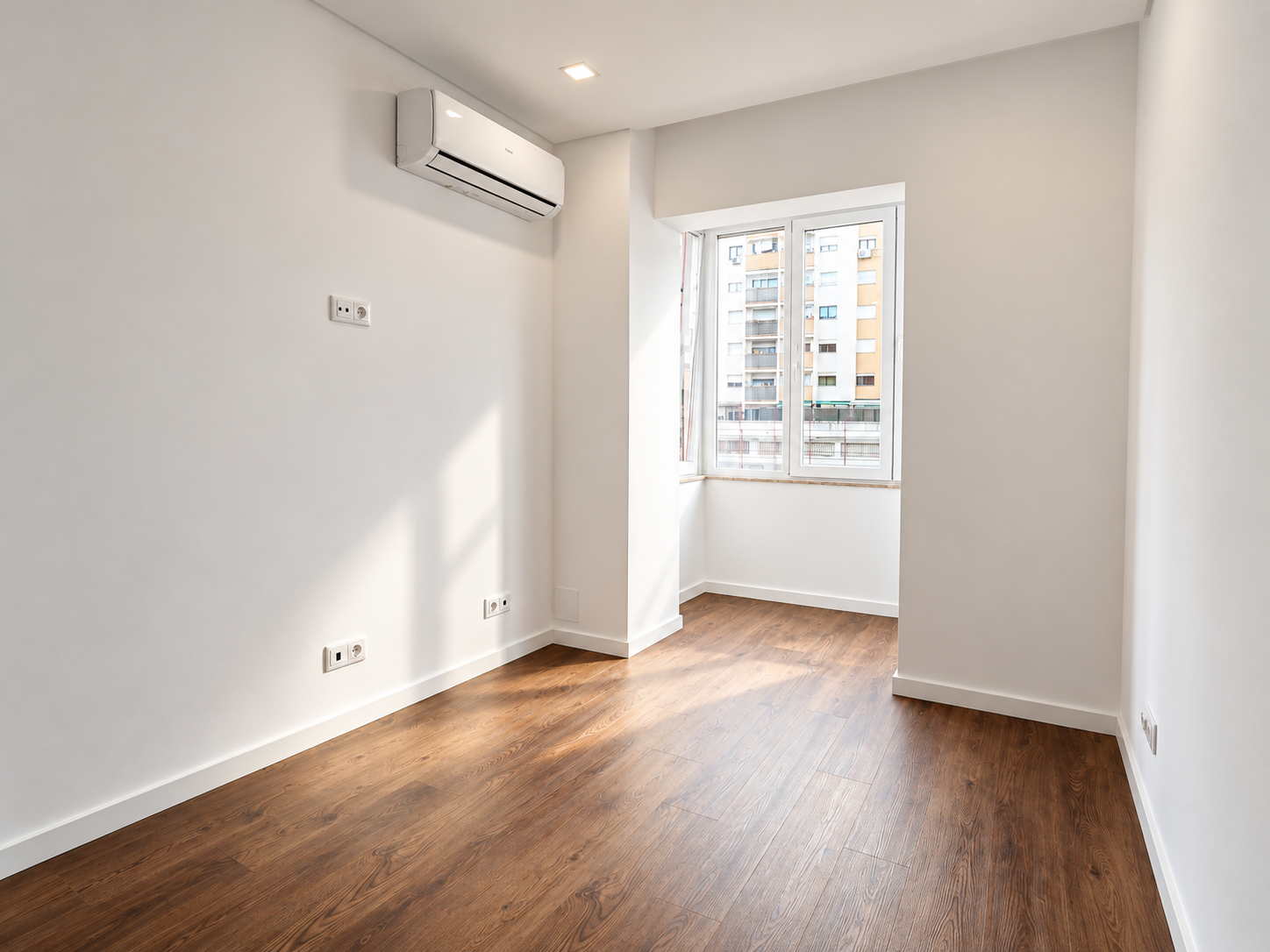 Empty bedroom with a white-framed window, wood-effect flooring and air conditioning on the left-hand wall.