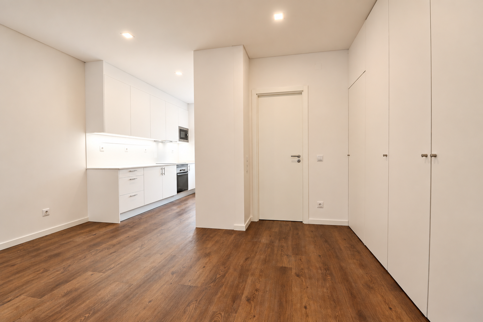 Living room with wood-effect flooring, full-height white cupboards and an open kitchen in the background.