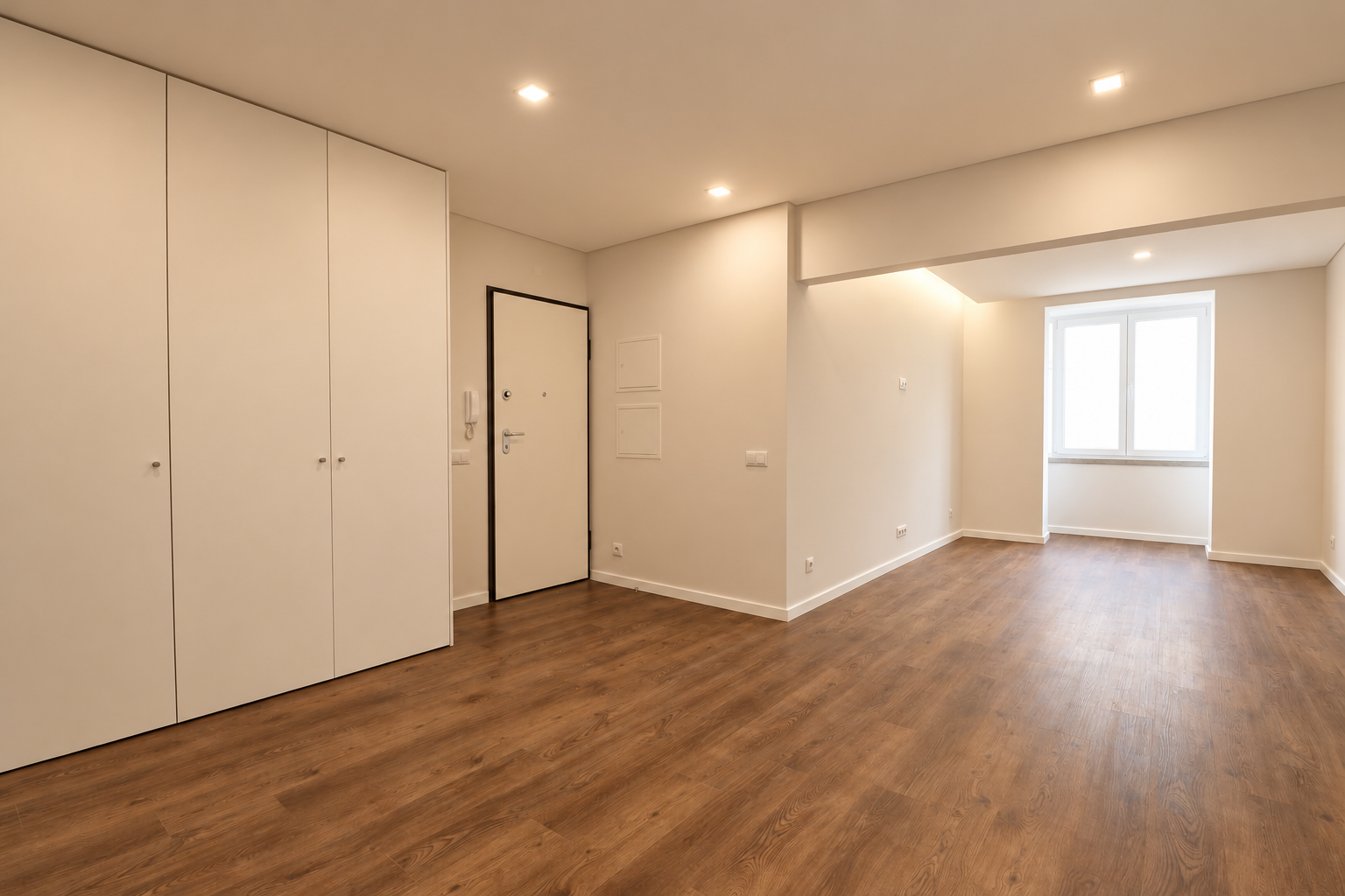 Empty living room with white cupboards on the left, the entrance door and a window at the far end.