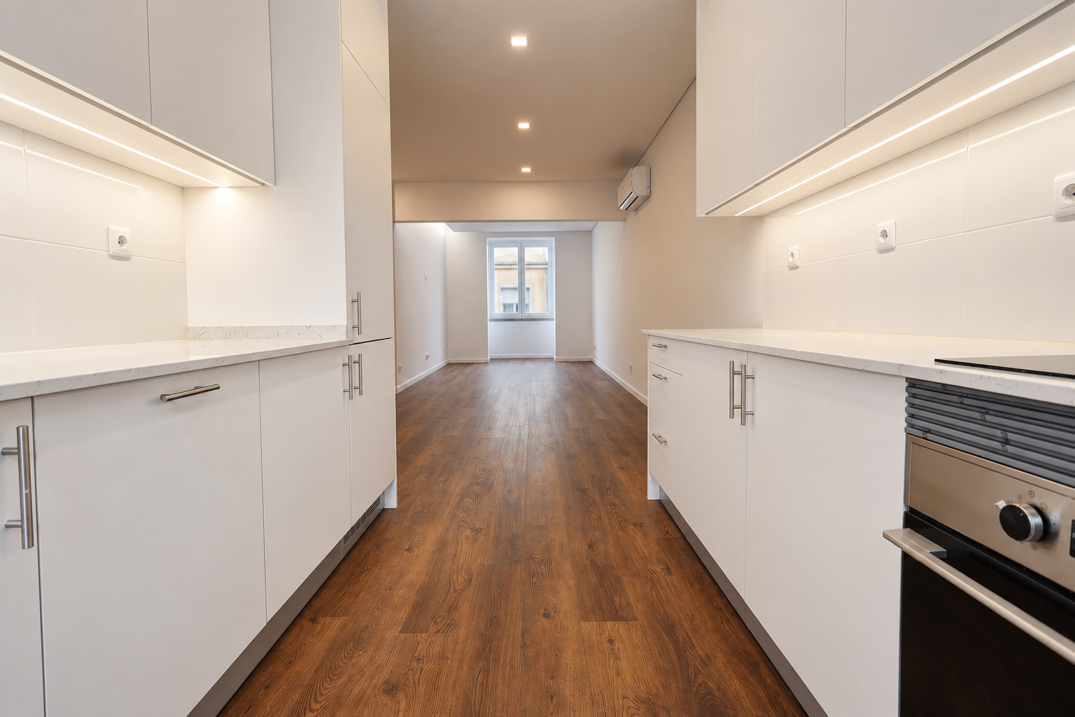 Kitchen with parallel worktops, white cabinets and an oven on the right, opening onto a living room with a window and air conditioning.