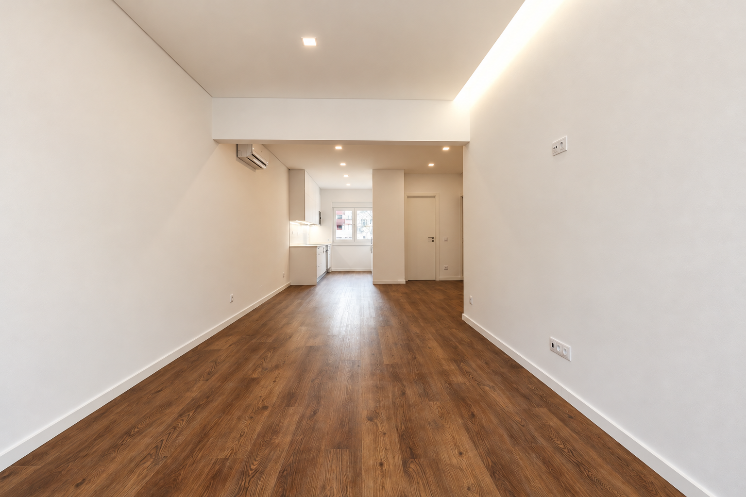 Long living room with wood-effect flooring, indirect ceiling lighting and a view towards the white kitchen and window at the far end.