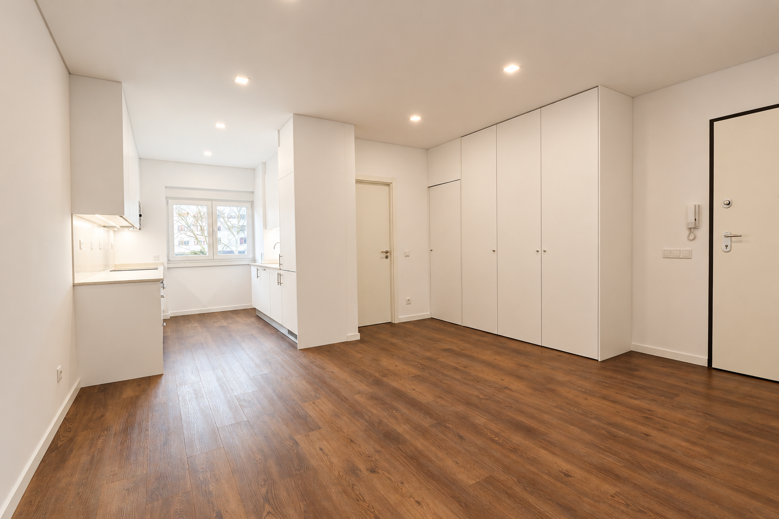 Empty living room with wood-effect flooring, a white kitchen beside the window and full-height cupboards next to the entrance door.